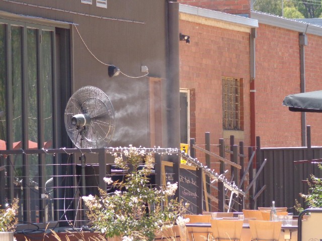 A fan blowing droplets of water across a cafe's outdoor seating area to keep it cool.